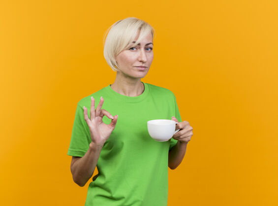 市民自信的中年金发斯拉夫女人端着一杯茶 看着前方做着“ok”的标志 隔离在黄色的墙上 留着复制空间表情女人茶