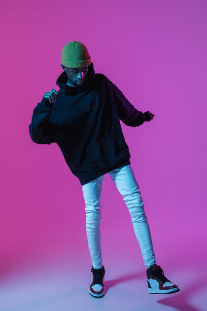 动作年轻时尚的男人穿着现代街头风格的服装 孤零零地站在霓虹灯下的斜墙上 非洲裔美国时尚模特在看音乐人表演衣服渐变名人