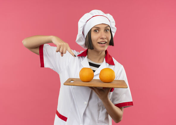 板子身着厨师制服的快乐年轻女厨师手拿并指着插着橘子的砧板 上面是粉红色的拿着女指着