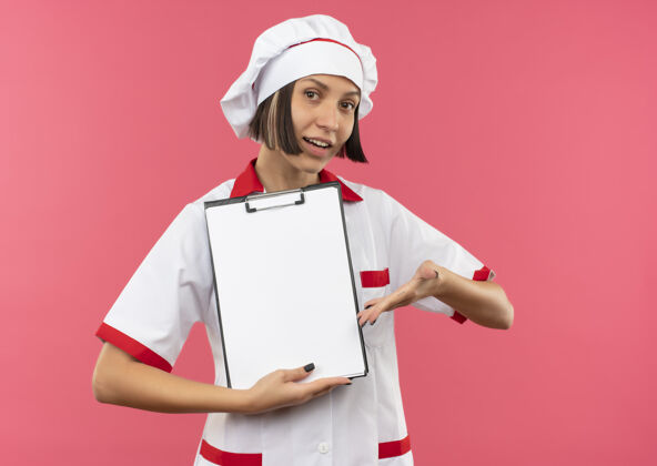 女身着厨师制服的年轻女厨师手拿并指着粉色背景上的剪贴板指着年轻请