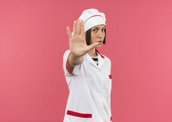 停止穿着厨师制服的年轻女厨师站在粉色背景下 对着镜头做手势女性年轻厨师