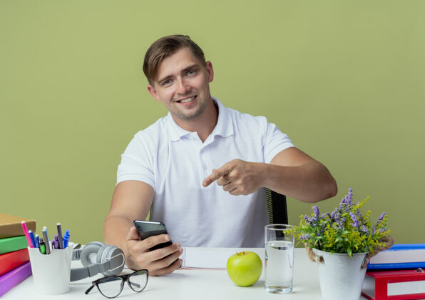 电话面带微笑的年轻帅哥坐在书桌旁 手里拿着学习用具 指着橄榄绿上孤立的电话橄榄学生学校