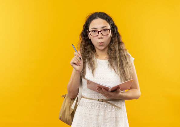 背令人印象深刻的年轻漂亮的女学生戴着眼镜 背着提包拿着钢笔和笔记本隔离在黄色拿着印象笔