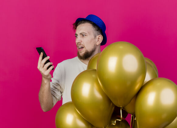 人一个年轻英俊的斯拉夫人 戴着派对帽 站在气球后面 拿着手机看着粉红色的墙上衣服抱着手机
