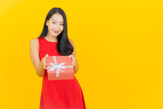 亚洲人肖像美丽的亚洲年轻女子微笑与红色礼品盒在黄色的墙壁节日年轻人庆祝