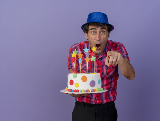 人印象深刻的中年党人戴着党的帽子拿着生日蛋糕看着和指着前面孤立的紫色墙壁与复制空间穿着人拿着