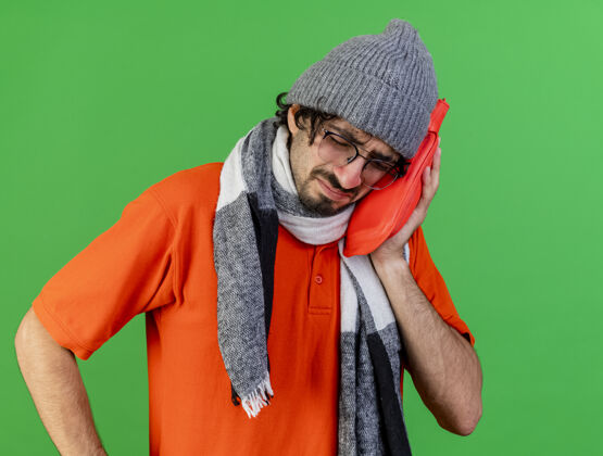 疾病愁眉不展的年轻病夫戴着眼镜 戴着冬天的帽子和围巾 手里拿着热水袋 闭着眼睛抚摸着脸 隔离在绿色的墙上帽子穿着姿势
