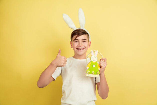 有趣竖起大拇指装饰白人男孩作为一个复活节兔子在黄色的工作室背景复活节快乐的问候美丽的男模人类情感的概念 面部表情 节日复制空间时尚创意表情