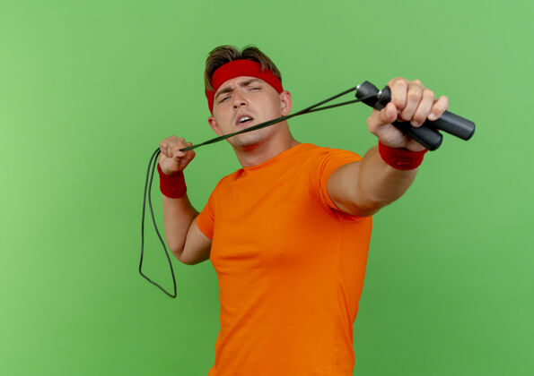 伸展戴着头带和腕带的年轻帅气的运动型男人 在绿色背景下 对着镜头拉着和拉着的跳绳腕带绳子运动
