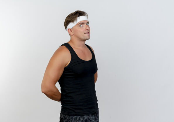 运动印象深刻的年轻英俊的运动男子戴着头带和腕带站在侧视图把双手放在背后隔离在白色头带站立佩戴