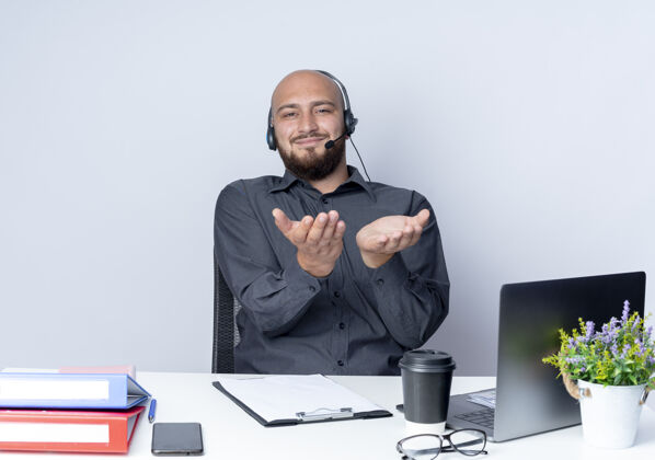 显示高兴的年轻秃头呼叫中心男子戴着耳机坐在办公桌旁 工作工具显示空手对着隔离在白色背景上的摄像头秃头穿工作