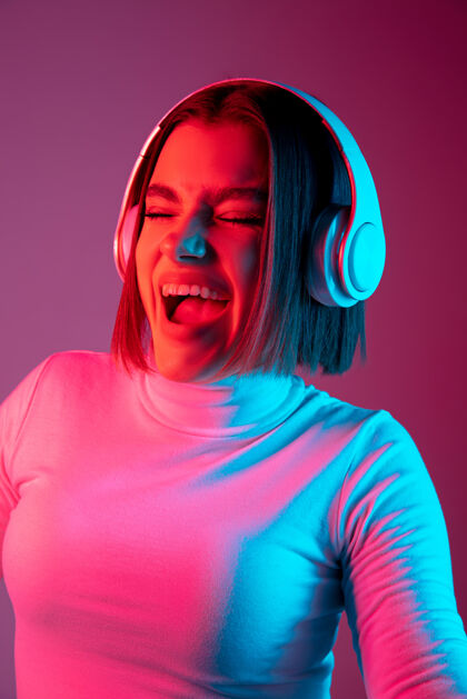 街道音乐流行霓虹灯下粉红色工作室背景上的白种女人肖像戴着耳机的漂亮女模特人类情感的概念 面部表情 销售 广告 时尚手霓虹灯城市