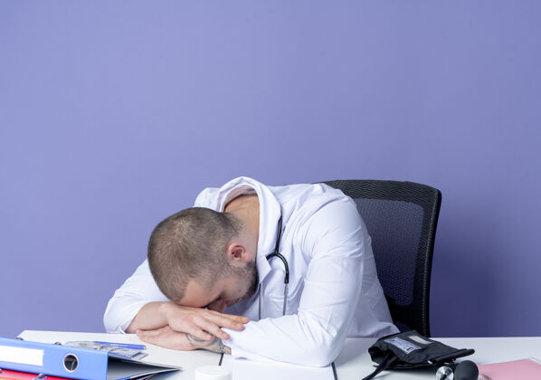 年轻疲惫的年轻男医生穿着医用长袍和听诊器坐在办公桌旁 手放在办公桌上 头放在手上 隔离在紫色背景上长袍坐着工具