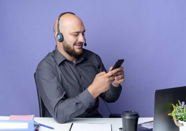 中心高兴的年轻秃头呼叫中心男子戴着耳机坐在办公桌前 用他的手机在紫色的背景上孤立的工作工具请戴秃头电话