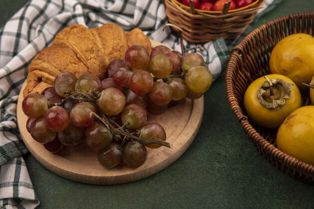 水平顶视图葡萄在一个木制的厨房板上的格子布与牛角面包柿子水果桶上的绿色背景葡萄羊角面包柿子