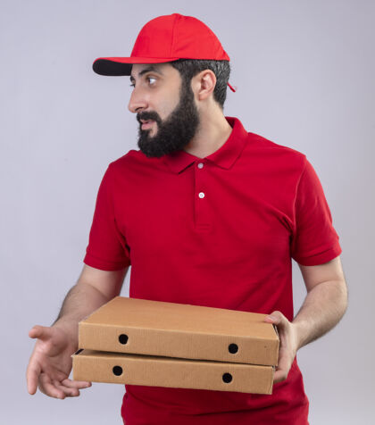帽子年轻英俊的白人送货员身穿红色制服 头戴鸭舌帽 手里拿着披萨盒 看着白色背景上孤立的一面送货年轻盒子