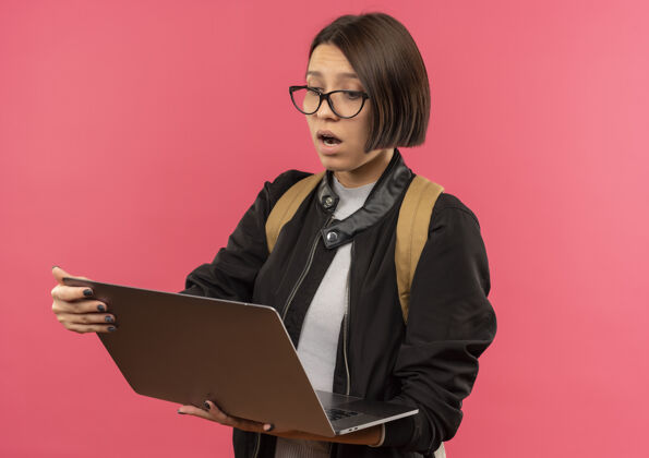 抱印象深刻的年轻女学生戴着眼镜 背着书包 看着笔记本电脑上孤立的粉红色粉红色眼镜穿