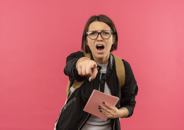 粉红色印象深刻的年轻女学生戴着眼镜 背着手袋拿着便签本指着前面孤立的粉红色眼镜年轻学生