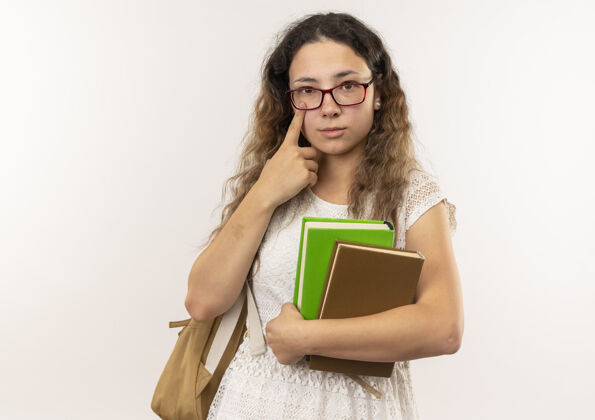 穿年轻漂亮的女学生戴着眼镜 背着书包 拿着书 手指放在眼睛下面 看着隔离在白色背景上的相机 还有复印空间手指眼睛抱