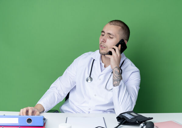 电话年轻的男医生穿着医用长袍和听诊器坐在办公桌旁 手里拿着工作工具 一边打电话 一边低头看着桌子 背景是绿色的坐着医疗穿