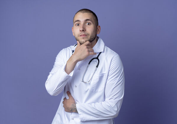 佩戴年轻的男医生穿着医用长袍 脖子上戴着听诊器 手放在肚子上 触摸紫色的下巴印象医学医生