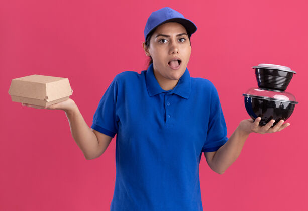年轻一个穿着制服 戴着帽子 拿着食物容器 双手分开放在粉红色墙上的年轻送货女孩惊讶不已蔓延容器食物