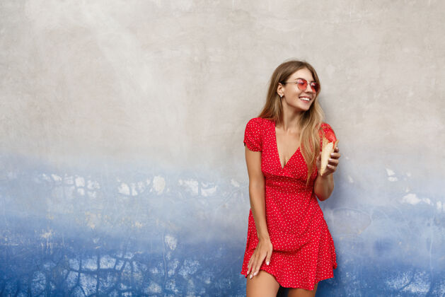乐趣戴着红色太阳镜 穿着夏装的时尚都市女孩 在户外吃着冰淇淋 倚在水泥墙上望着远方太阳镜女孩冰淇淋