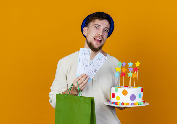 举行令人印象深刻的年轻英俊的斯拉夫党家伙戴着党的帽子拿着机票纸袋和生日蛋糕与明星看着相机隔离在橙色背景与复制空间复制票帽子