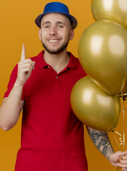 帽子面带微笑的年轻英俊的斯拉夫党人戴着党的帽子拿着气球看着摄像机指着孤立的橙色背景派对年轻人指着