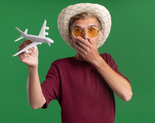 掩护一个穿着红衬衫戴着眼镜戴着帽子的年轻帅哥拿着玩具飞机捂着嘴 手被隔离在绿色的墙上嘴巴害怕穿