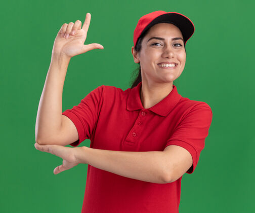 微笑微笑的年轻送货女孩穿着制服 戴着帽子 在绿色的墙上显示出的姿势穿表演手势
