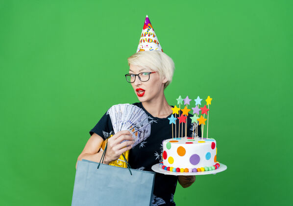 复制令人印象深刻的年轻金发派对女孩戴着眼镜和生日帽拿着生日蛋糕与明星金钱礼盒和纸袋看着相机隔离在绿色背景与复制空间蛋糕钱金发