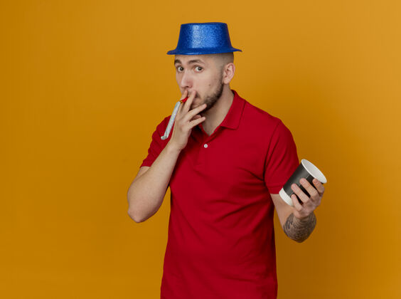空间令人印象深刻的年轻英俊的斯拉夫党的家伙戴着党的帽子看着相机举行党吹嘴与塑料咖啡杯在手上孤立的橙色背景与复制空间橙色家伙塑料