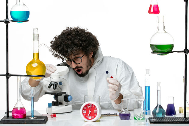 实验室外套正面图身穿特殊套装的年轻男性科学家拿着样品 在白墙上使用显微镜视图化学药剂师