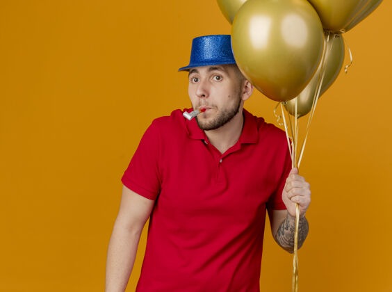橙色令人印象深刻的年轻英俊的斯拉夫党家伙戴着党帽子拿着气球看着一边举行党吹在嘴里孤立的橙色背景与复制空间帽子斯拉夫人气球