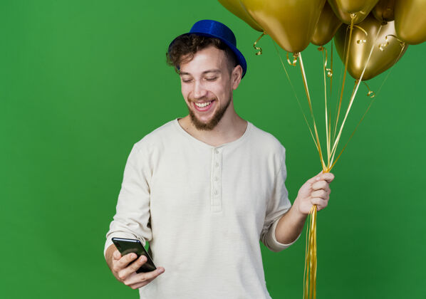 复制快乐的年轻英俊的斯拉夫党人戴着党的帽子拿着气球和手机看着手机在绿色背景与复制空间隔离手机拿着帽子