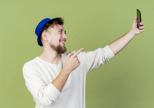 年轻快乐的年轻帅哥斯拉夫党家伙戴着党的帽子自拍指着手机孤立在橄榄绿的背景斯拉夫人派对自拍