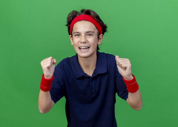绿色快乐的年轻帅气的运动男孩戴着头带和护腕 戴着牙套 看着相机在绿色背景上做着“是”的手势运动欢乐背带