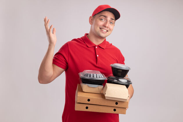 容器面带微笑的年轻送货员穿着制服 戴着帽子 拿着食物容器放在披萨盒上 手被隔离在白墙上穿传播食物