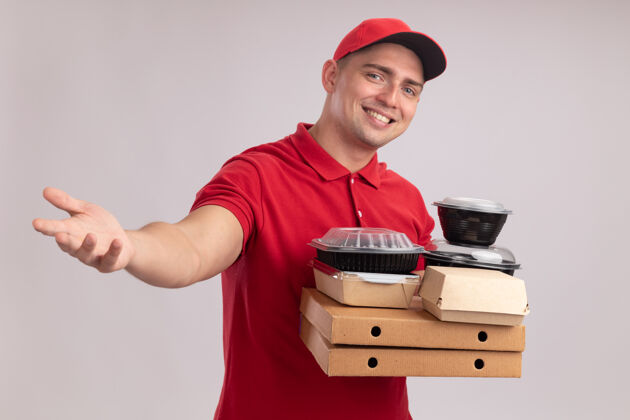 年轻人微笑的年轻送货员身穿制服 戴着帽子 拿着比萨饼盒上的食品容器 手伸向隔离在白墙上的摄像机容器制服微笑