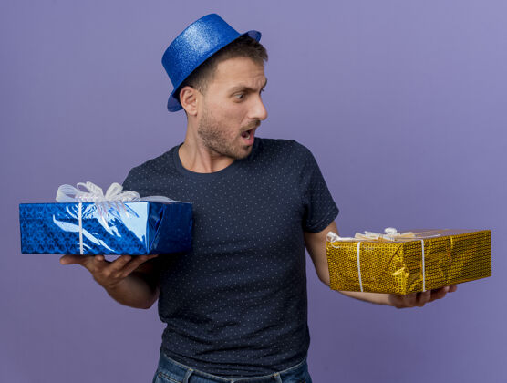 蓝色震惊英俊的白人男子戴着蓝色帽子举行 并期待着礼品盒孤立的紫色背景与复制空间盒子持有空格