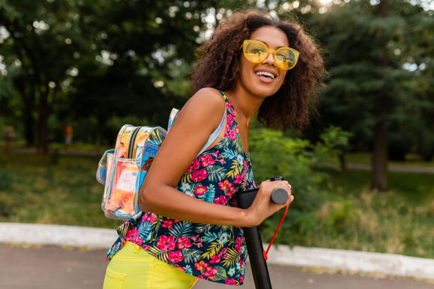 街道年轻时尚的黑人女子在公园里玩得很开心骑着电动脚踏车在夏天的时尚风格 五颜六色的时髦服装 穿着背包和黄色太阳镜埃斯库特摩托车环保