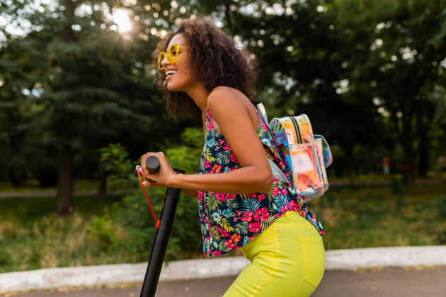 积极年轻时尚的黑人女子在公园里玩得很开心骑着电动脚踏车在夏天的时尚风格 五颜六色的时髦服装 穿着背包和黄色太阳镜埃斯库特车辆环保