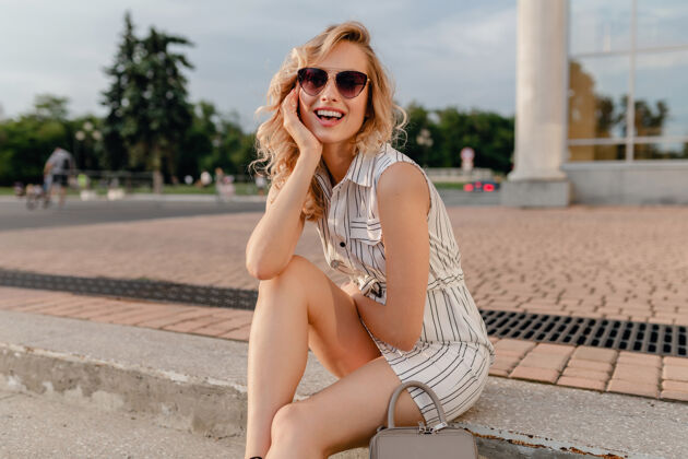 年轻年轻迷人可爱时尚的金发女郎坐在城市街道上夏日时尚的白色棉质连衣裙戴着墨镜女性城市城市