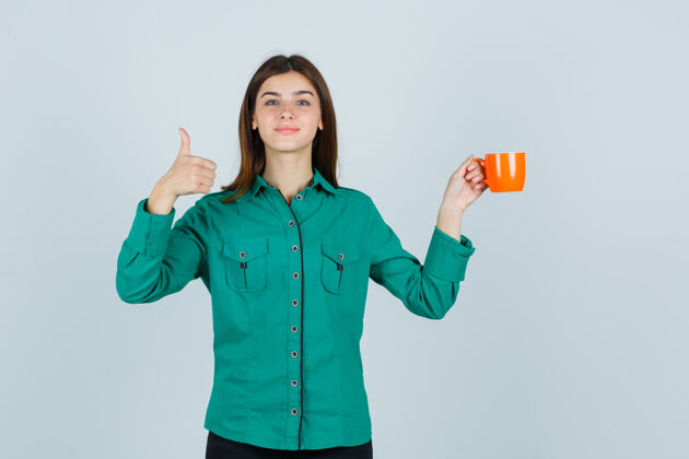 女人一位年轻的女士拿着一杯橘色的茶 同时在衬衫上竖起大拇指 看上去很高兴年轻而衬衫