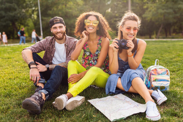 青春快乐的年轻朋友们一起坐在公园里 男女一起玩 带着相机旅行 聊天 微笑女人黄色多种族