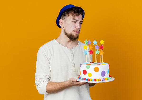 抱着令人印象深刻的年轻英俊的斯拉夫党人戴着党的帽子举行生日蛋糕与明星看直橙色背景与复制空间孤立直年轻人小伙子