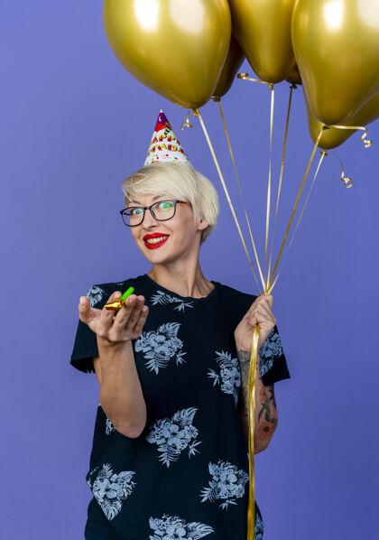 眼镜微笑的金发派对女郎戴着眼镜 戴着生日帽 手里拿着气球和派对吹风机 看着紫色背景上孤立的摄像机生日气球年轻