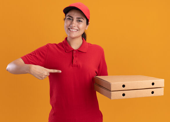 制服微笑着的年轻送货女孩穿着制服 戴着帽子 指着隔离在橙色墙上的比萨饼盒送货点女孩