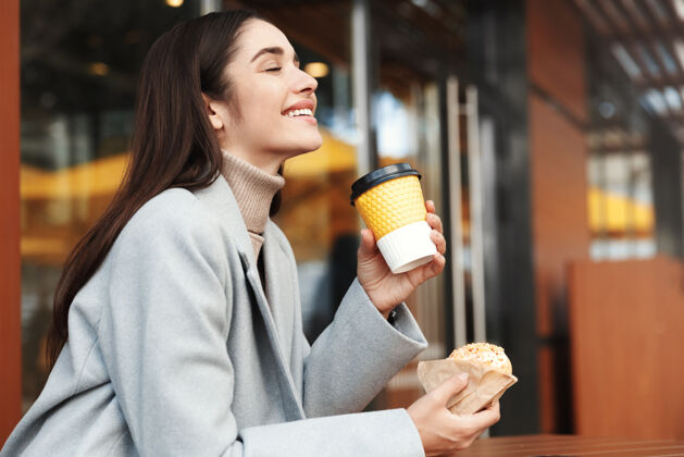 女商人穿着灰色外套的快乐的年轻女人在咖啡店吃甜甜圈放松抱着早餐